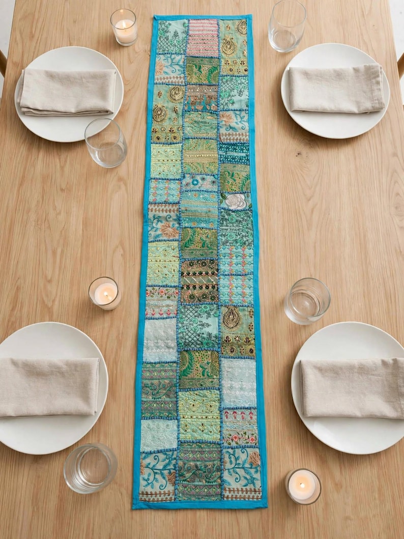 Puede incluir: Un camino de mesa rectangular con un dise&ntilde;o de patchwork de varios cuadrados de tela en tonos verdes y azulados con intrincados bordados, abalorios y detalles en hilo met&aacute;lico, acabado con un borde liso de color azul brillante.