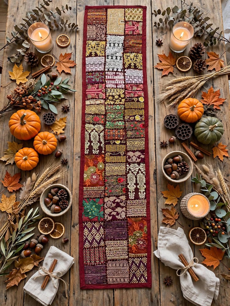 May include: A rectangular table runner featuring a patchwork design of various embroidered and beaded fabric squares in shades of burgundy, gold, and green, centered on a wooden surface surrounded by autumn-themed decorations.