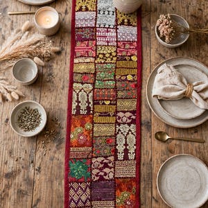 May include: A long, rectangular table runner featuring a patchwork design of various embroidered fabric squares in shades of red, gold, green, and purple, bordered by a solid deep red fabric trim, laid flat on a rustic wooden table.