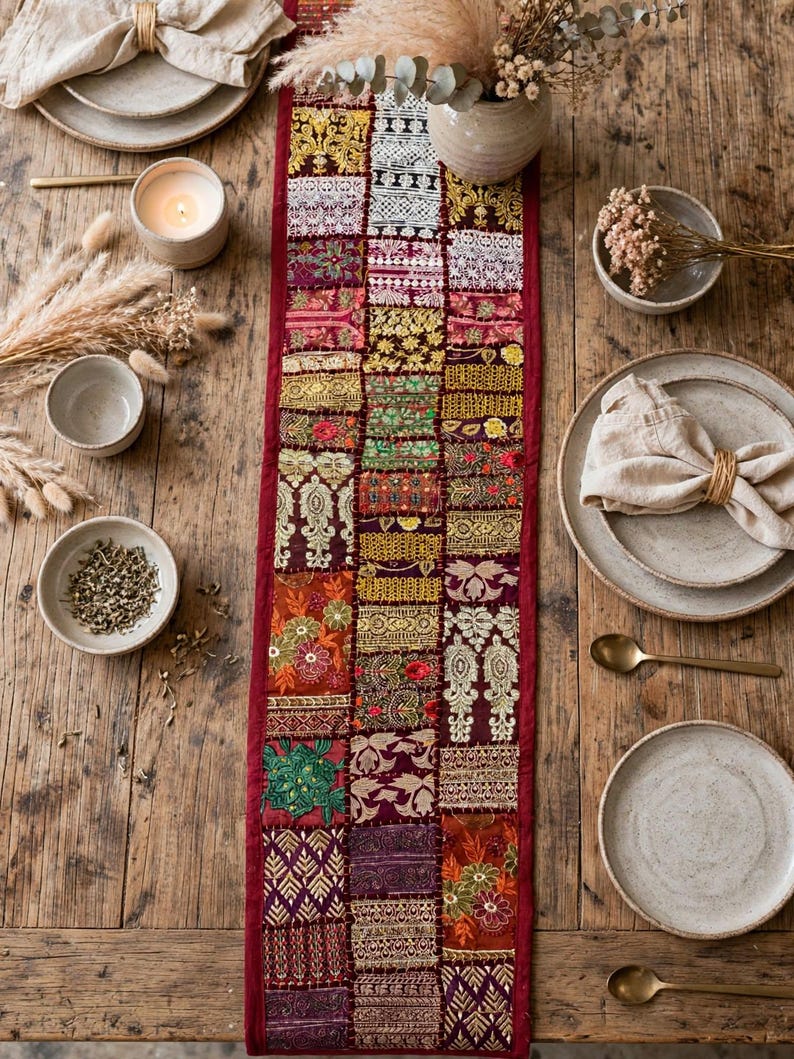 May include: A long, rectangular table runner featuring a patchwork design of various embroidered fabric squares in shades of red, gold, green, and purple, bordered by a solid deep red fabric trim, laid flat on a rustic wooden table.