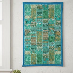 Puede incluir: Un tapiz de pared rectangular con un diseño de patchwork. La pieza presenta una variedad de telas verdes y turquesas en un patrón de cuadrícula, enmarcado por un borde azul liso. Las telas tienen diferentes texturas y patrones.