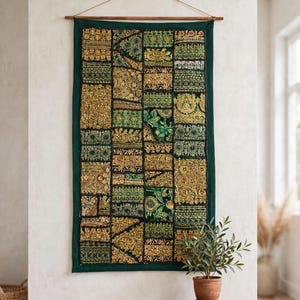 Puede incluir: Un tapiz rectangular que presenta un mosaico de paneles de tela en verde oscuro y dorado. El tejido muestra bordados intrincados, detalles en hilo metálico y adornos de lentejuelas dispuestos en un patrón de cuadrícula, suspendido de una varilla de madera.