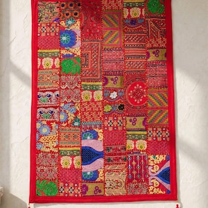 May include: A rectangular wall hanging featuring a vibrant patchwork design of various red, blue, green, and yellow fabric squares. The piece is bordered in solid red fabric and includes intricate embroidery, sequins, and two red corner tassels.
