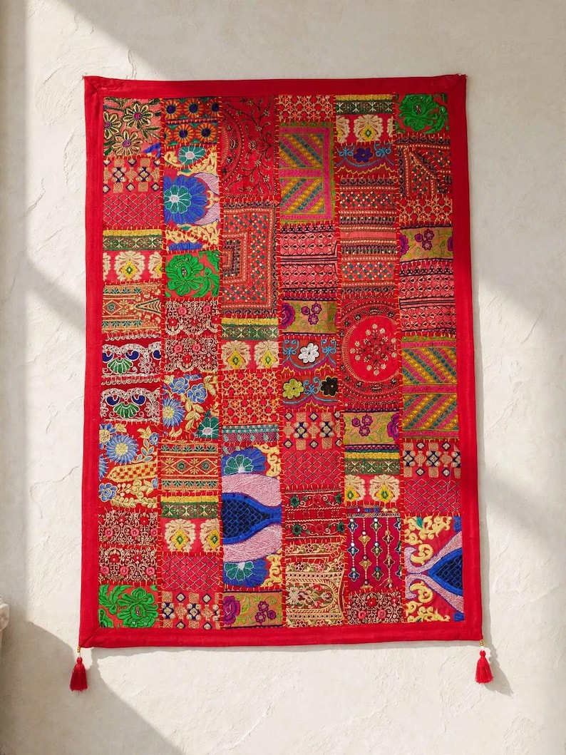 May include: A rectangular wall hanging featuring a vibrant patchwork design of various red, blue, green, and yellow fabric squares. The piece is bordered in solid red fabric and includes intricate embroidery, sequins, and two red corner tassels.