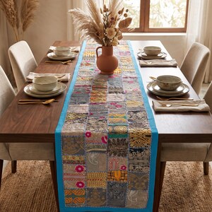 May include: A rectangular table runner featuring a patchwork design of various fabric squares in shades of gray, gold, and pink, bordered by a bright turquoise trim. The runner is placed centrally on a dark wood dining table with place settings.