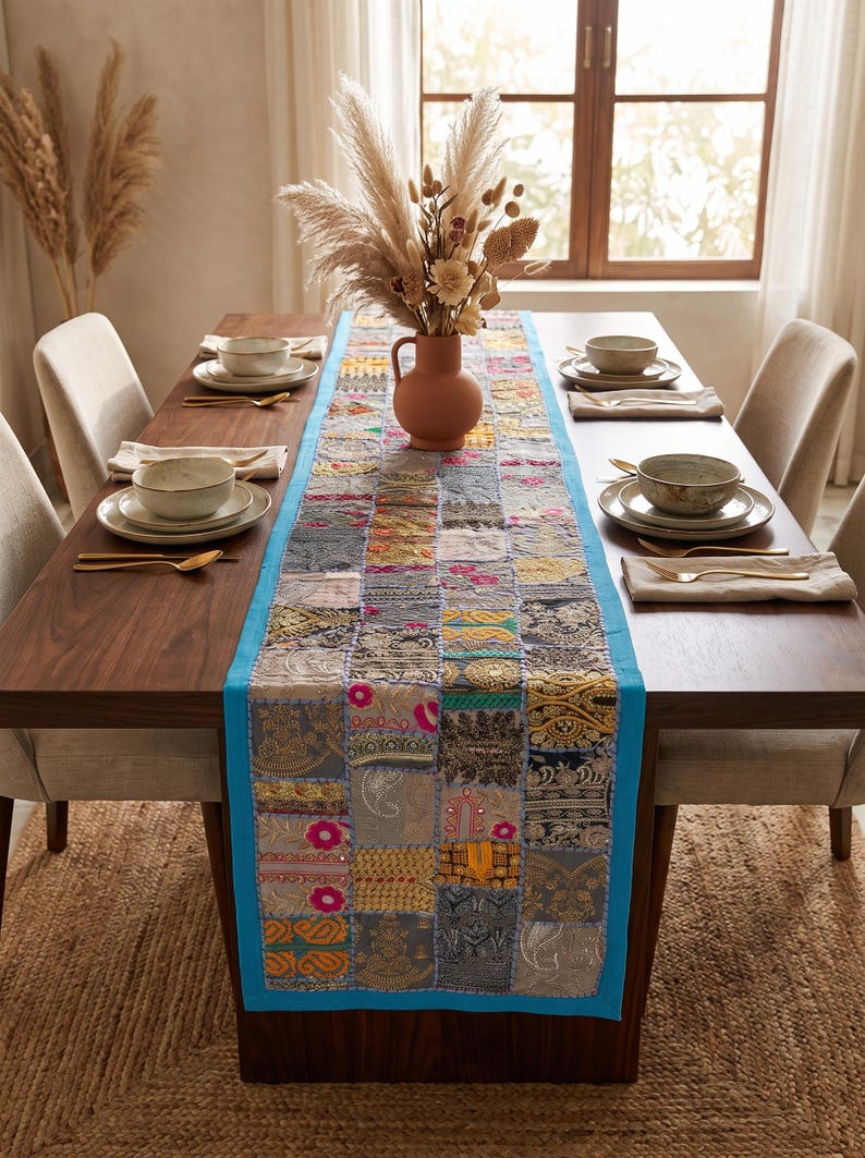 May include: A rectangular table runner featuring a patchwork design of various fabric squares in shades of gray, gold, and pink, bordered by a bright turquoise trim. The runner is placed centrally on a dark wood dining table with place settings.