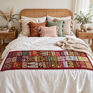 May include: A rectangular bed runner featuring a patchwork design of various embroidered fabrics in shades of red, gold, green, and purple. The runner is laid across a white bedspread, showcasing intricate patterns and textures in each fabric square.