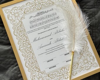 May include: A framed marriage certificate with gold accents and ornate detailing. The certificate features Arabic script and the names Muhammad Abduli and Aminah Abduti. A white feather quill with a gold-colored handle rests on the document.