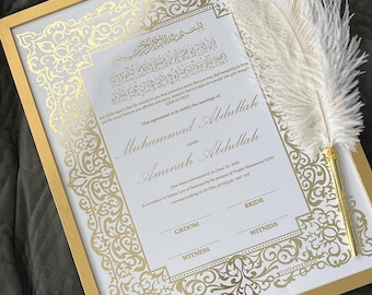 May include: A framed marriage certificate with gold accents and ornate detailing. The certificate includes names and dates, with a decorative feather quill resting on the frame. The frame is gold and the background is white.