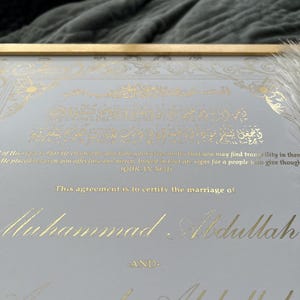 May include: Framed marriage certificate with gold foil calligraphy on a light gray background. The text includes Arabic script and the names Muhammad Abdullah and Aminah Abdullah. The frame is gold.