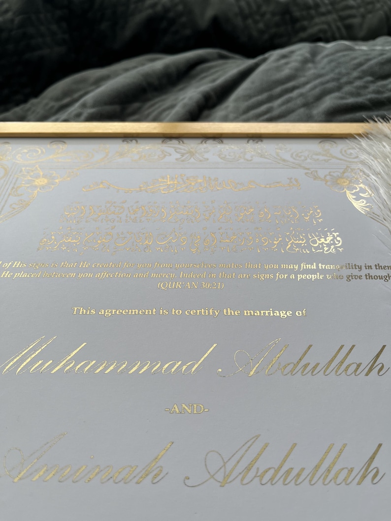 May include: Framed marriage certificate with gold foil calligraphy on a light gray background. The text includes Arabic script and the names Muhammad Abdullah and Aminah Abdullah. The frame is gold.