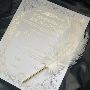 May include: A white wedding certificate with gold foil calligraphy and floral border. A white feather quill pen with a gold-colored metal base rests on the certificate. The certificate includes text in Arabic and English.