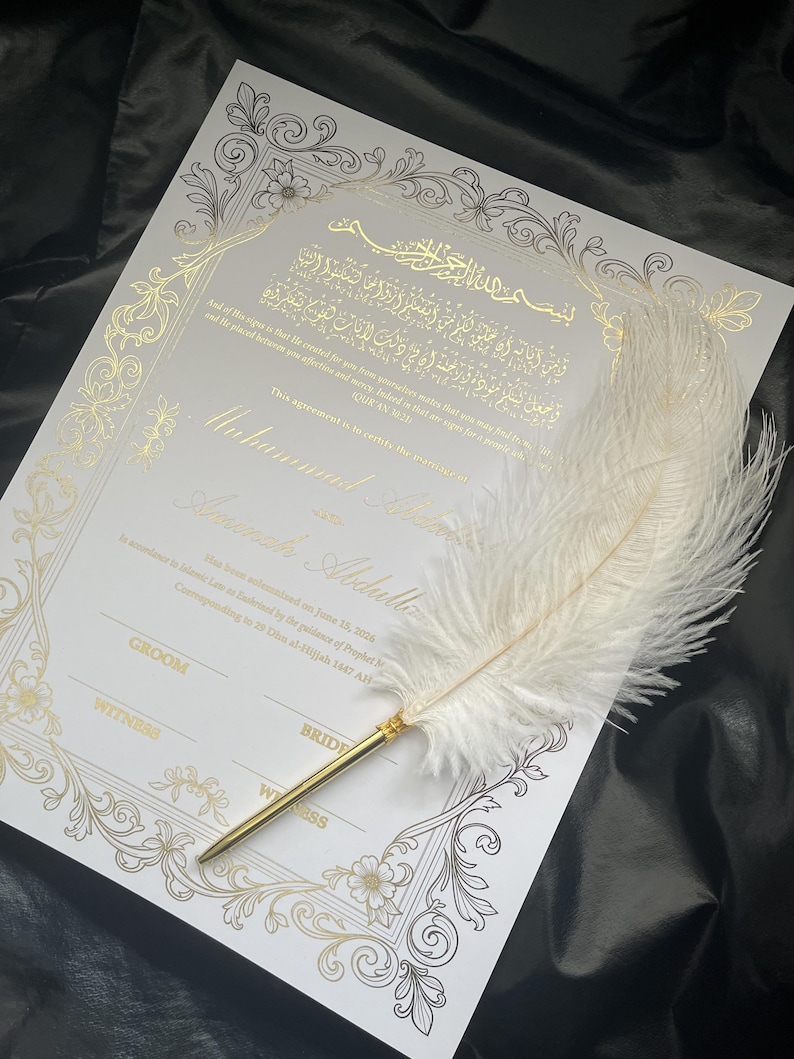 May include: A white wedding certificate with gold foil calligraphy and floral border. A white feather quill pen with a gold-colored metal base rests on the certificate. The certificate includes text in Arabic and English.