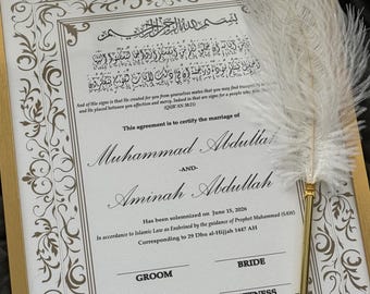 May include: A framed marriage certificate with ornate brown border details and text in English and Arabic. A white feather quill with a gold-colored metal base rests on the frame. The certificate includes the names Muhammad Abdullah and Aminah Abdullah.