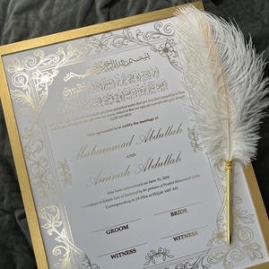Peut inclure: Un certificat de mariage encadré avec des accents dorés et des motifs floraux ornés. Le certificat comprend une écriture arabe et les noms Muhammad Abdullah et Aminah Abdullah. Un stylo plume blanc repose sur le cadre.