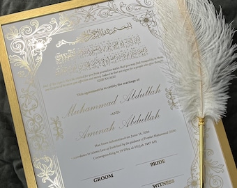 May include: A framed marriage certificate with gold accents and ornate floral designs. The certificate includes Arabic script and the names Muhammad Abdullah and Aminah Abdullah. A white feather pen rests on the frame.