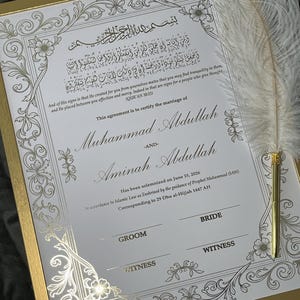 May include: A framed marriage certificate with gold accents and ornate floral designs. The document features Arabic script and English text, including names and a date. A white feather quill rests on the frame, adding an elegant touch.