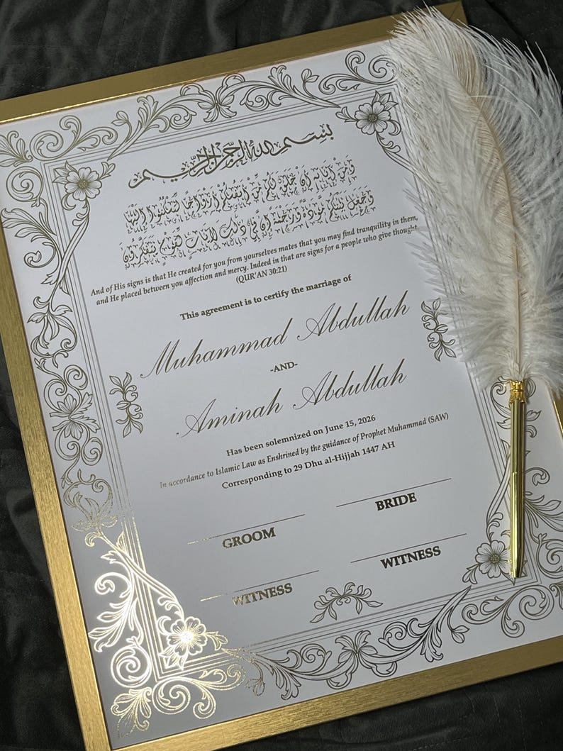 May include: A framed marriage certificate with gold accents and ornate floral designs. The document features Arabic script and English text, including names and a date. A white feather quill rests on the frame, adding an elegant touch.
