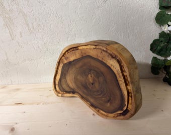May include: A handcrafted, irregularly shaped wooden cutting board. The board features a rich, natural wood grain with varying shades of brown and a lighter-colored border. The wood's unique shape and grain patterns are visible.