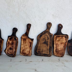 Può includere: Sei taglieri rustici in legno con manici, ognuno con una forma unica e bordi scuri e carbonizzati. I taglieri hanno una finitura naturale a venature del legno, perfetti per la cucina.