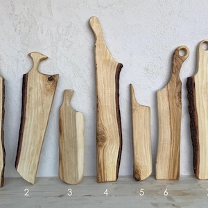 Può includere: Una collezione di sette taglieri in legno con bordi naturali. Le tavole variano in forma e dimensione, mostrando la venatura del legno e i bordi naturali della corteccia. Ogni tagliere è unico, perfetto per servire o preparare il cibo.