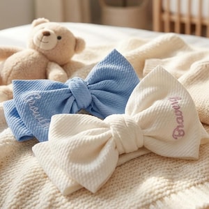 Puede incluir: Dos diademas de lazo para bebé, una azul claro y otra blanquecina, con texto bordado. Un osito de peluche beige, un mordedor de madera y un jersey color crema también están en la imagen.