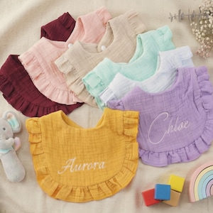Puede incluir: Una colección de baberos para bebés con volantes en varios colores pastel, incluyendo amarillo, morado y rosa. Los baberos tienen nombres bordados y están dispuestos con un sonajero de elefante gris, bloques de madera y un juguete en forma de arcoíris. Los baberos son de tela suave y texturizada.