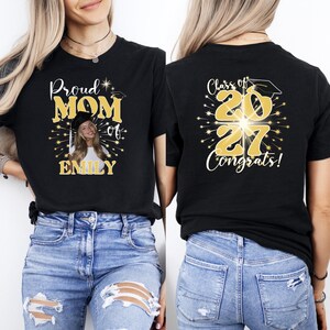 May include: A black t-shirt featuring a custom graphic design. The front displays the text Proud MOM of EMILY with a photo of a graduate. The back features the text Class of 2027 Congrats! in gold lettering with a graduation cap and firework accents.