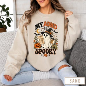 May include: A sand-colored crewneck sweatshirt featuring a graphic print with the text "MY ADHD IS SPOOKY". The design includes a ghost wearing a witch hat, holding a coffee cup and a to-do list, surrounded by pumpkins, bats, a moon, and a brain icon.