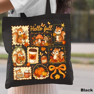 May include: A black tote bag featuring a colorful autumn-themed graphic with the text Hello fall. The design includes illustrations of squirrels, mushrooms, a coffee cup, a candle, pumpkins, and a bow, all surrounded by fall leaves and butterflies.