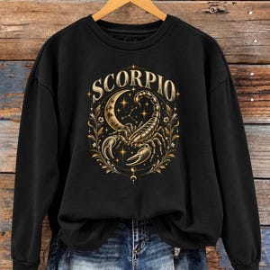 May include: A black long-sleeve sweatshirt featuring a gold-toned graphic print of a scorpion, a crescent moon, stars, and floral elements. The text SCORPIO is printed in a curved font above the central illustration.