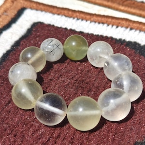 May include: A bracelet composed of translucent, spherical beads in clear, yellow, and green hues. Some beads feature internal inclusions. The bracelet is presented on a maroon surface.