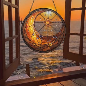 May include: A round stained glass window depicting a crescent moon and a starburst design, with a chain for hanging. The window is in shades of gold, orange, and brown, with clear and blue accents. The window is hanging in front of an open window overlooking the ocean.