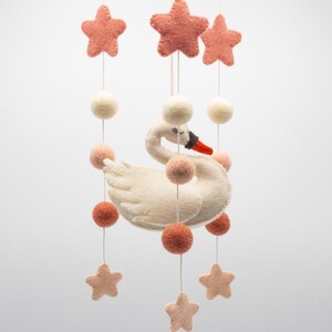 May include: A nursery mobile with a white felt swan, black beak and orange accents, and pink and cream felt stars and balls. The mobile is suspended by white strings, creating a whimsical decoration. The mobile is designed for a baby's room.