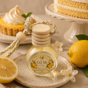 May include: A small, round glass car air freshener bottle with a light wooden cap and a braided rope hanger. The label features yellow stripes, lemon illustrations, and the text EAT ME, Lemon Creme, Almond, Vanilla, and The Mad Candler.