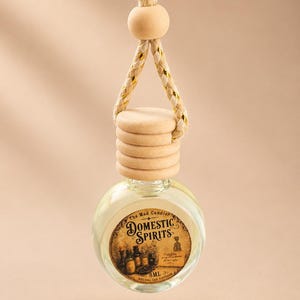 May include: A round glass car diffuser bottle with a light wooden cap and a braided rope hanger featuring gold metallic threads. The circular label reads The Mad Candler Domestic Spirits and 8ML Natural Car Diffuser with vintage-style graphics.