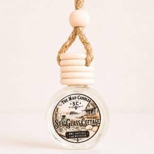 May include: A clear glass car diffuser bottle with a wooden cap and a beige rope hanger. The label features a coastal cottage illustration with the text The Mad Candler, SC, Sea Glass Cottage, and 8ml natural car diffuser printed on the front.