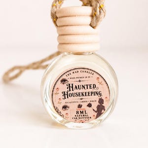 May include: A clear glass car diffuser bottle with a light wood cap and a braided jute hanging cord. The label reads: THE MAD CANDLER, HAND-POURED IN SC, HAUNTED HOUSEKEEPING, EUCALYPTUS AMBER BIRCH, 8ML NATURAL CAR DIFFUSER, with eye and silhouette graphics.