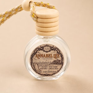May include: A clear glass 8mL car diffuser bottle with a light wood cap and a braided cord. The label features the text Annabel Lee, Car Diffuser, In a kingdom by the, Sea Mist, Rose Storm, Bergamot, Amkoss Drift, and 8mL Car Diffuser.