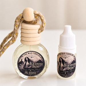 May include: A clear glass 8ml car diffuser bottle with a wooden cap and a braided rope, accompanied by a small refill bottle. Both feature labels with the text The Mad Candler, Blackwater Bride, and an illustration of a woman by the water.
