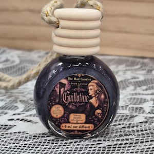 May include: A round glass car diffuser bottle filled with dark shimmering liquid, topped with a ribbed wooden cap and a braided cord. The label features the text The Mad Candler, Guillotine, Paris 1791, and 8 ml car diffuser.