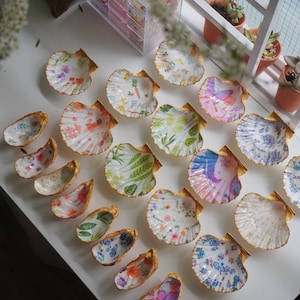 Op de afbeelding: Een verzameling decoratieve schelpen met goud geschilderde randen. De schelpen zijn beschilderd met diverse bloemen-, botanische en grillige ontwerpen. Sommige schelpen zijn open, andere gesloten, wat een gevarieerde weergave creëert.