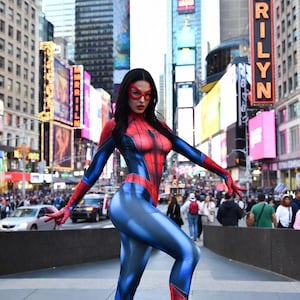May include: A person in a Spider-Man costume poses in Times Square, New York City. The costume is red and blue with a web design on the chest. The person wears a red mask and black high-heeled boots. The background includes city buildings and billboards.