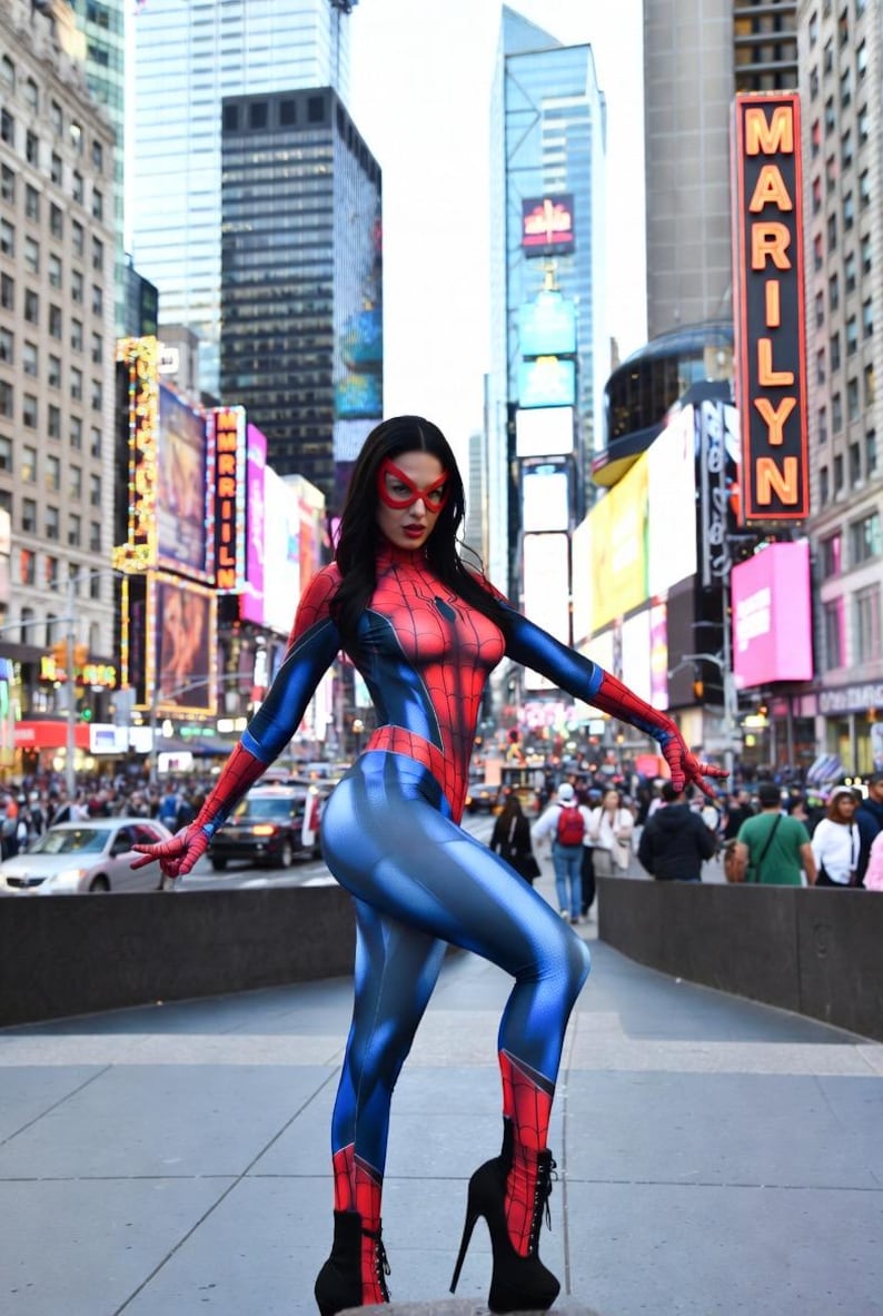 May include: A person in a Spider-Man costume poses in Times Square, New York City. The costume is red and blue with a web design on the chest. The person wears a red mask and black high-heeled boots. The background includes city buildings and billboards.