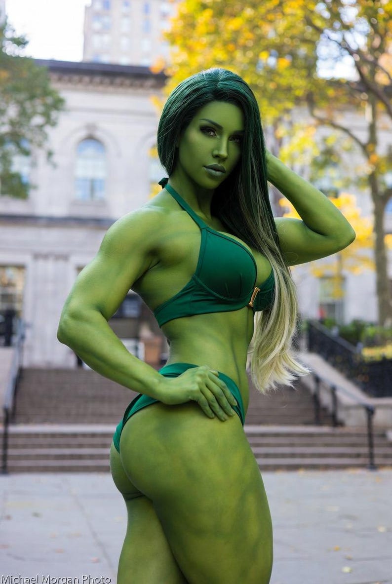 May include: A person with green skin and hair, wearing a dark green bikini. The person is standing outdoors with a building and trees in the background. The person is posing with one hand on their head and the other on their hip.