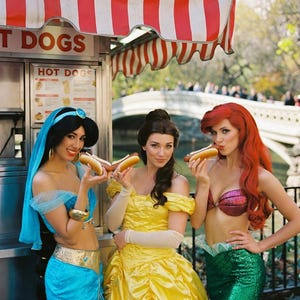 May include: Three women dressed as Disney princesses pose with hot dogs in front of a food stand. The stand has a red and white striped awning and the words "HOT DOGS" are visible. The princesses are dressed in blue, yellow, and green outfits.
