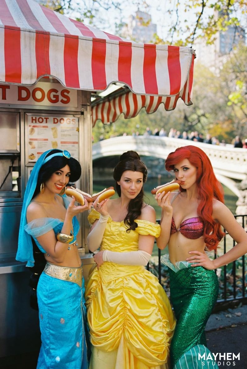May include: Three women dressed as Disney princesses pose with hot dogs in front of a food stand. The stand has a red and white striped awning and the words "HOT DOGS" are visible. The princesses are dressed in blue, yellow, and green outfits.
