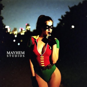 May include: A person wearing a red, green, and yellow bodysuit with a black mask and gloves. The bodysuit has a zipper down the front and a yellow belt. The background is dark with city lights and the text "MAYHEM STUDIOS".
