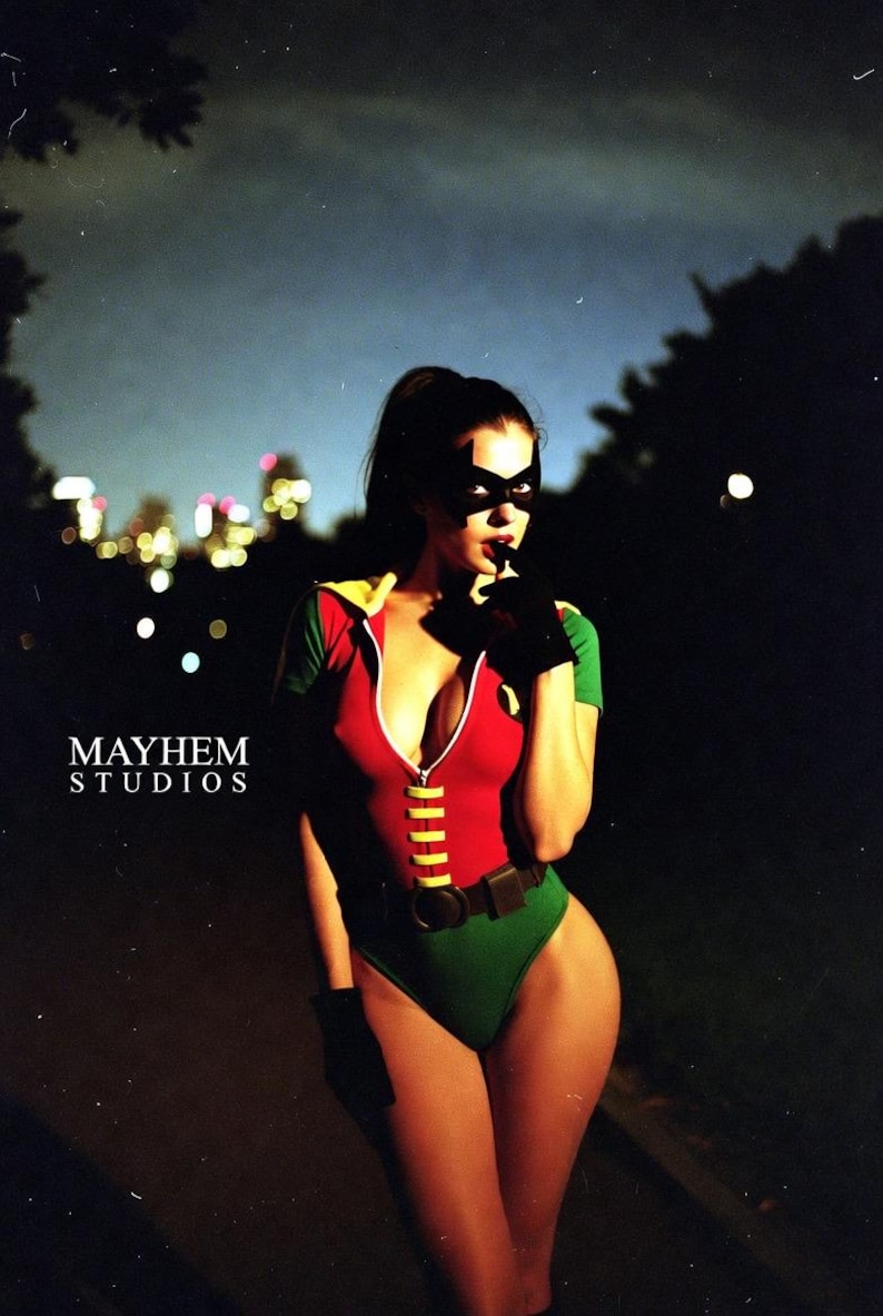 May include: A person wearing a red, green, and yellow bodysuit with a black mask and gloves. The bodysuit has a zipper down the front and a yellow belt. The background is dark with city lights and the text "MAYHEM STUDIOS".