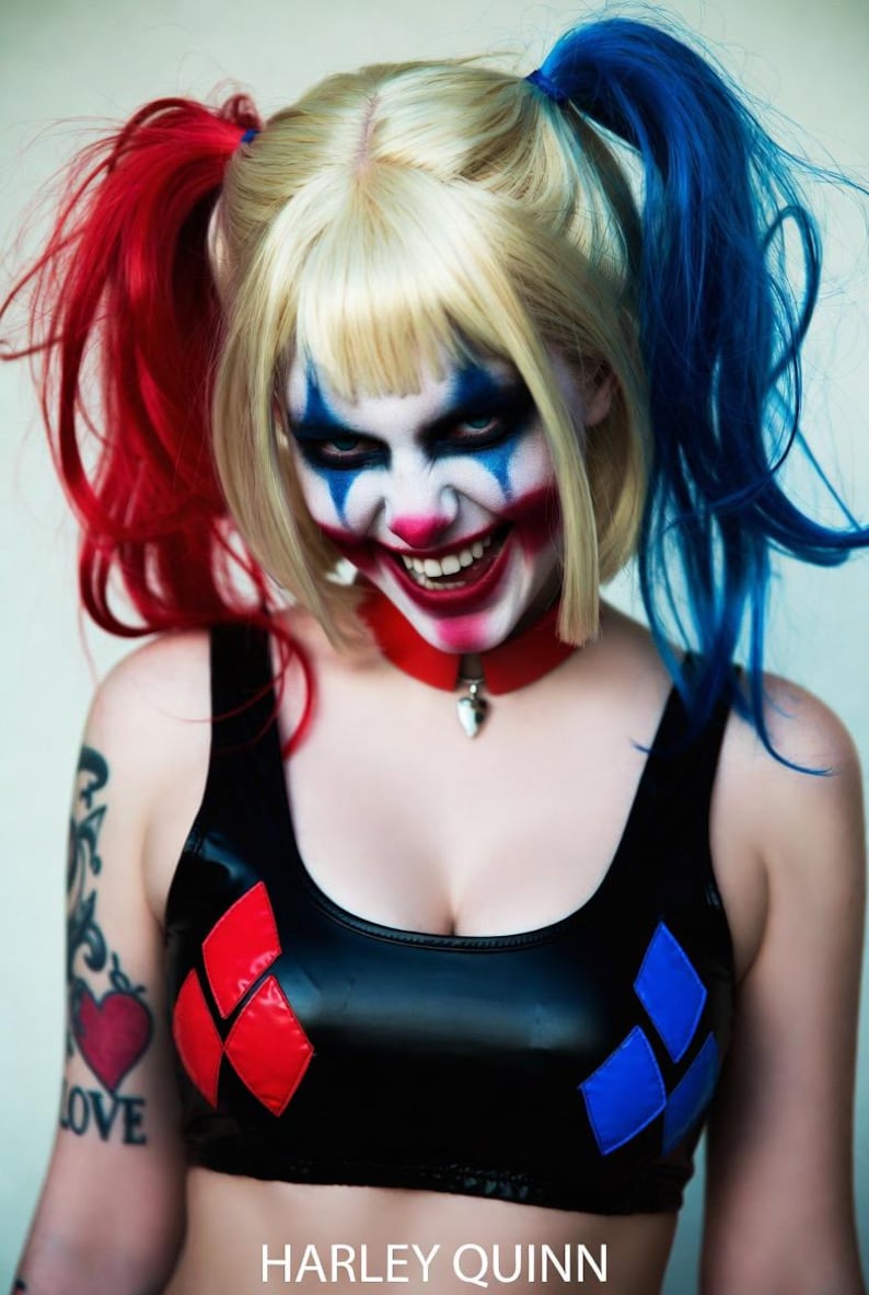 May include: A person with blonde hair styled in pigtails, one red and one blue. The person is wearing a black top with red and blue diamond shapes. The face is painted with a clown-like design, and the text "HARLEY QUINN" is visible.